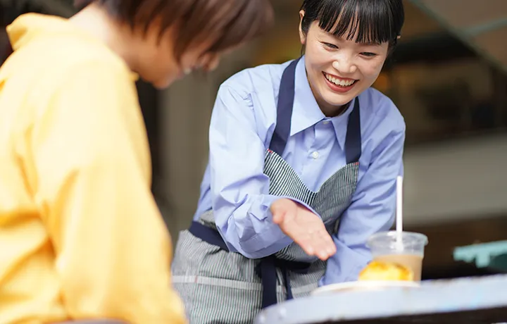 飲食店のカウンターで、笑顔の女性店員が客に手を差し出し、明るく接客している様子。
