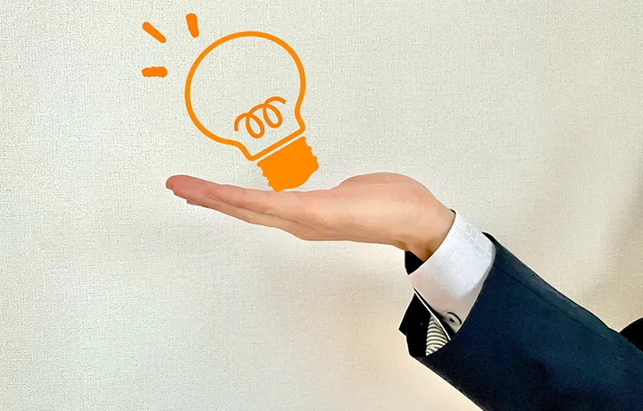 スーツ姿の人物が手のひらを差し出し、その上にオレンジ色の電球イラストが浮かんでいる。