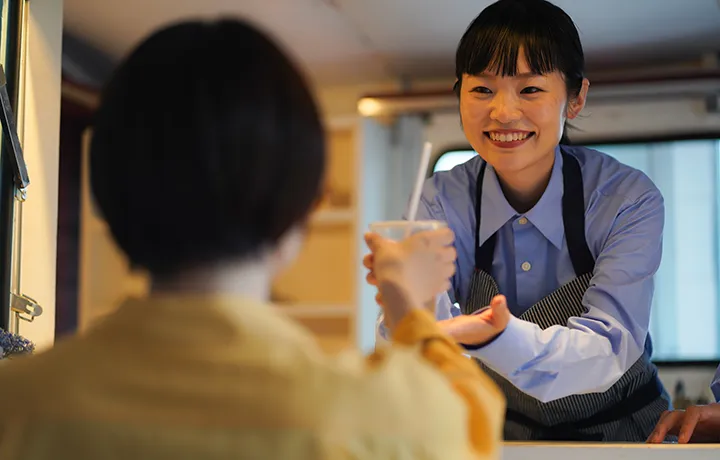 飲食店のカウンターで、笑顔の女性店員が客に飲み物の入ったカップを手渡している。